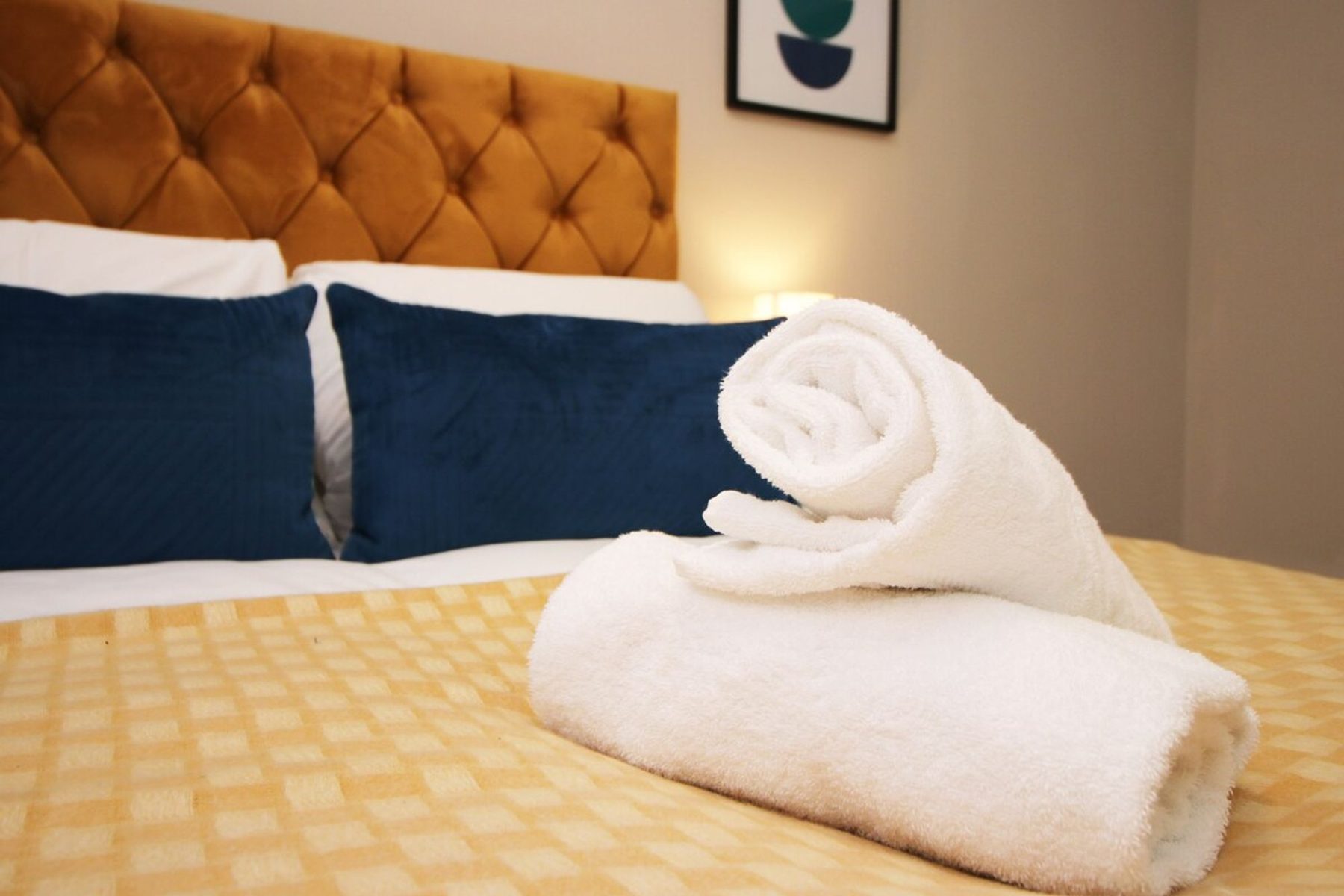 A folded white towel on a made bed with a mustard headboard in an EKLiving apartment